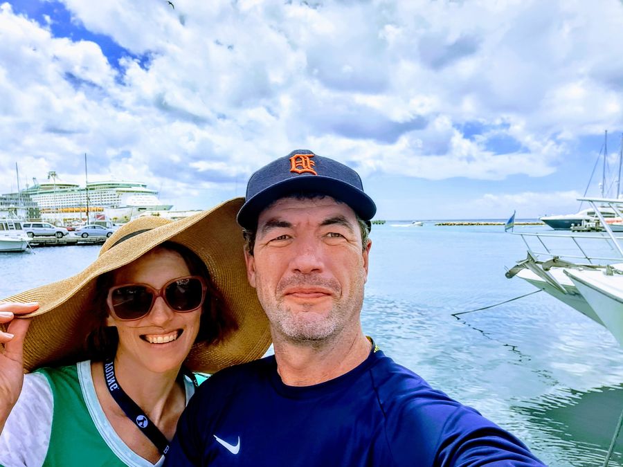 Ken and Lisa at Seaport Village Marina in Aruba, catamarans moored behind them and a cruise ship at the pier
